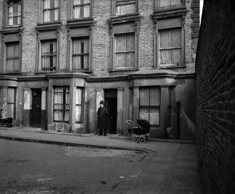 Black and white photo of Rillington Place in London, where multiple murders occurred. A police officer stands outside a house.
