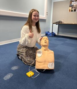Teenage girl giving a thumbs-up next to a CPR training dummy.