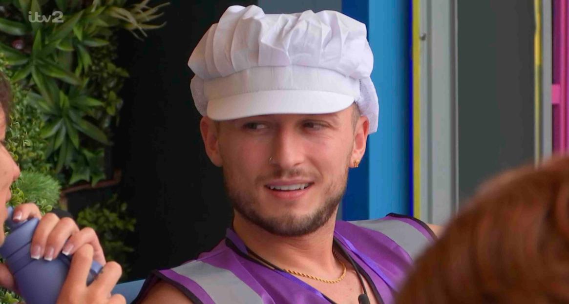 Sam wearing a chef's hat and purple vest, smiling while chatting with someone off-camera.