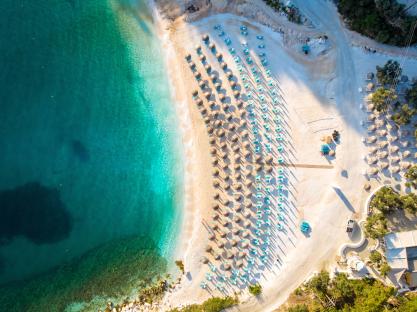 Aerial view of Marble Beach in Thasos, Greece, showing numerous umbrellas and beach chairs on the sand next to turquoise water.