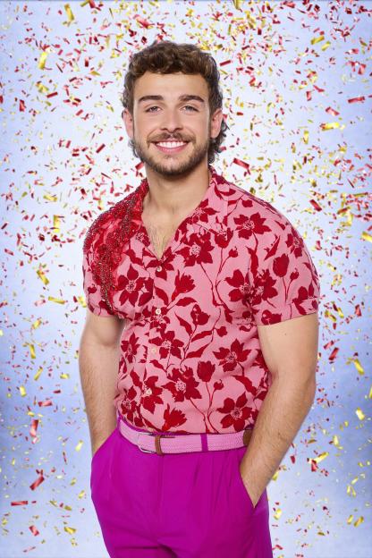 George Clarke smiling, dressed in a red floral shirt and bright pink trousers, with red and gold confetti falling around him.