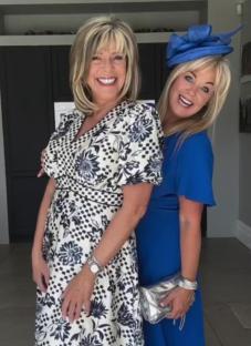 Two women at a garden party. One woman wears a black and white floral dress; the other wears a royal blue dress and a blue hat.