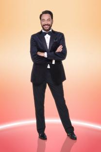 Craig Revel Horwood in a tuxedo, arms crossed, smiling.