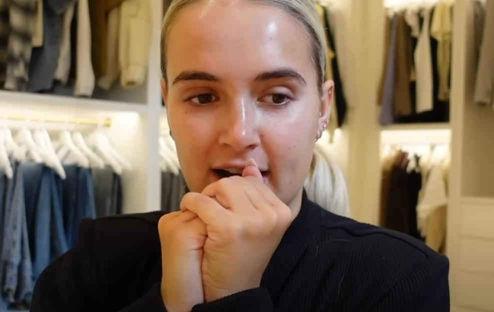 Screenshot of a woman in a closet, hands clasped in front of her.