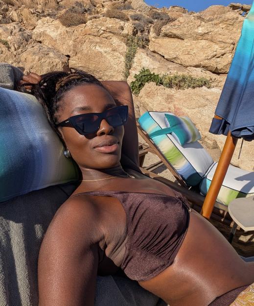 AJ Odudu wearing a brown bikini and sunglasses, reclining on a sun lounger with a rocky hillside in the background.