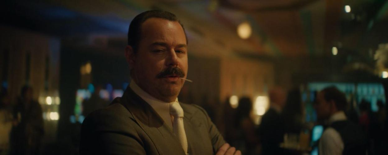 Man with a mustache wearing a suit, with a toothpick in his mouth, in a dimly lit bar.