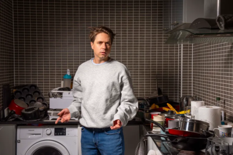 A man stands in a messy kitchen with dirty dishes.