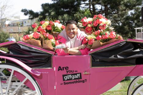 David Potts in a pink horse-drawn carriage with bouquets of flowers, holding a cake. The carriage is branded with