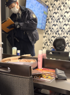 Screenshot of a police officer writing on a clipboard in a room, with toiletries and a person sitting in the background.