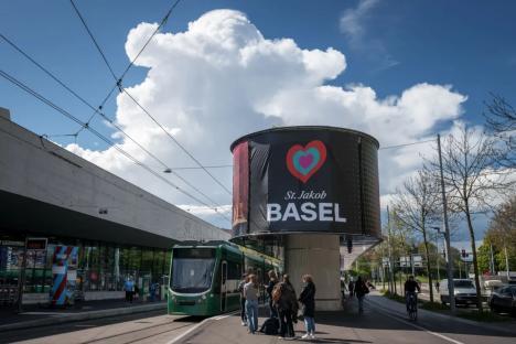 An image collage containing 1 images, Image 1 shows A tram stops near a large sign announcing Basel as the host city for the Eurovision Song Contest
