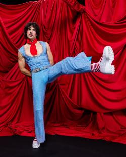 A performer in a sparkly blue jumpsuit and platform shoes poses against a red curtain.