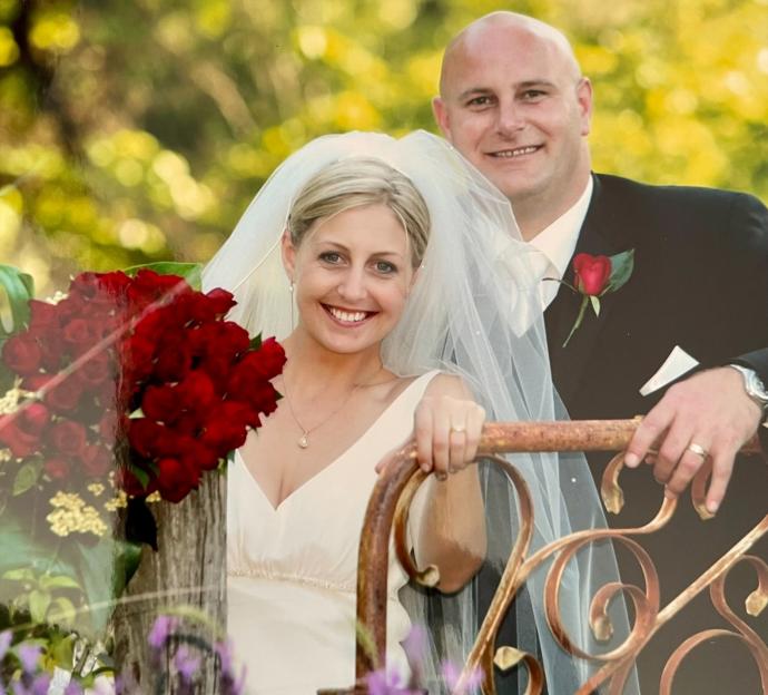 A newlywed couple, a woman in a white dress and veil holding a red rose bouquet, and a man in a black suit with a red rose boutonniere.