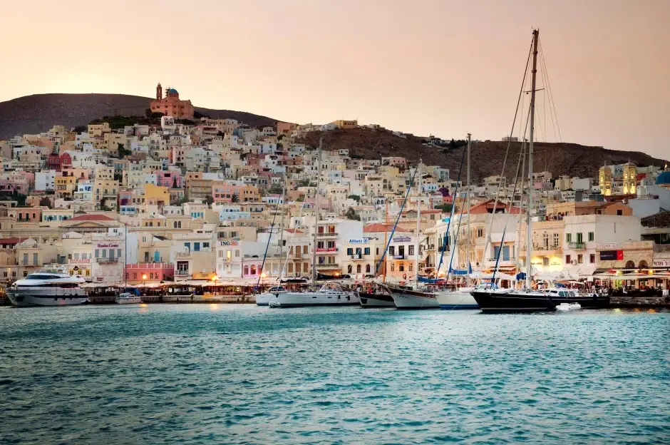 Coastal town of Syros, Greece, with buildings on a hill and boats docked in the foreground.