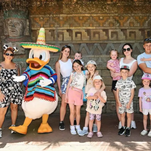 A large family group, including two adult women, a man, and several children, poses with Donald Duck dressed in a colorful serape and sombrero.