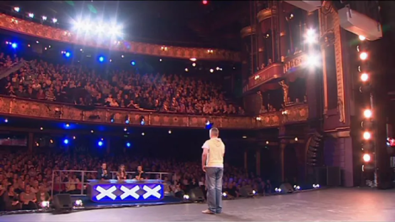 A man standing on a stage in front of a large audience during a televised talent show.