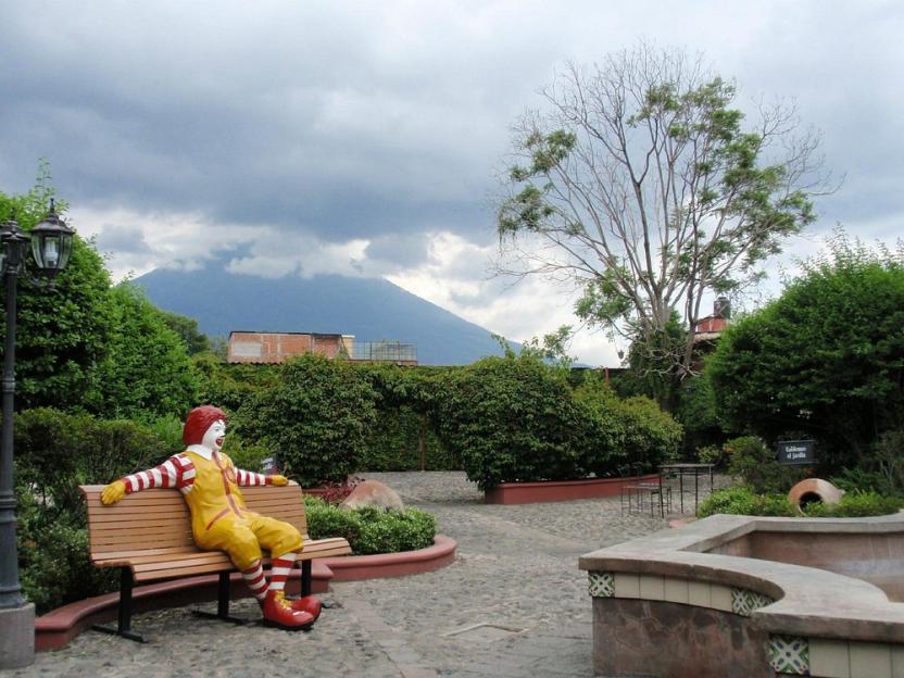 Ronald McDonald statue sitting on a bench in a courtyard with a volcano in the background.