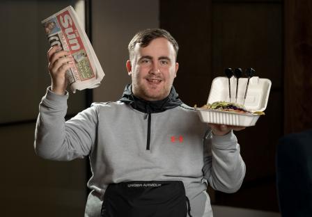 A young man holds a copy of The Sun newspaper and a kebab container with darts in it.