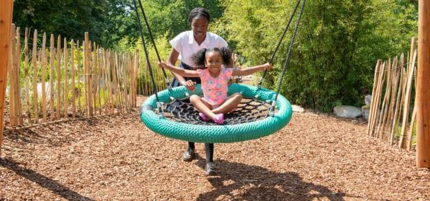 A young girl on a playground swing with a woman pushing her.