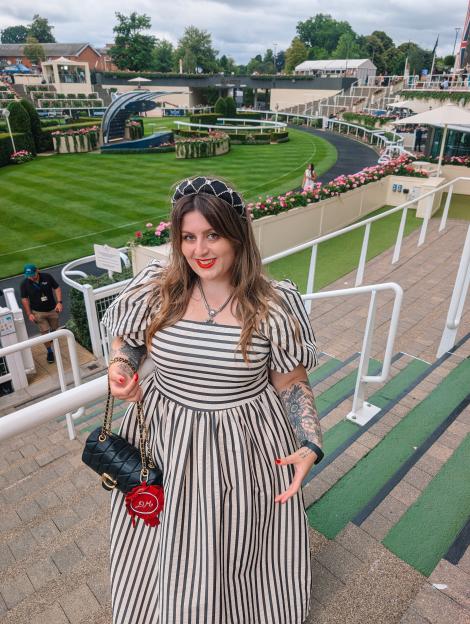 A woman in a black dress with red lipstick and a red necklace stands on a sidewalk in front of the Royal Albert Hall at night.