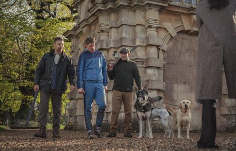 Three men with shotguns and two dogs stand before a woman.
