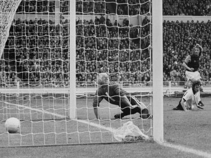 Black and white photo of a soccer goal, with the ball in the net, and players in the foreground.