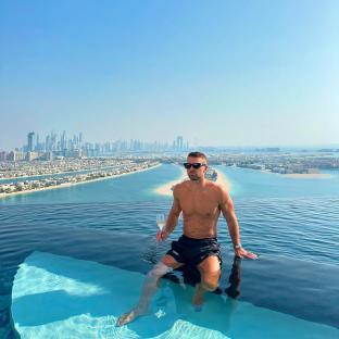 a shirtless man sits on the edge of an infinity pool holding a glass of champagne