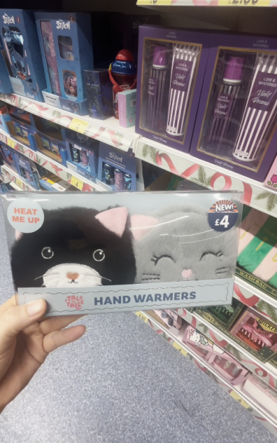 A hand holds a package of "Tale Tails Hand Warmers" depicting one black cat and one gray cat, each with a "HEAT ME UP" label and a "NEW! £4" sticker on the packaging.