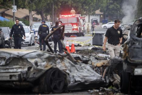 Aftermath of a plane crash in a residential neighborhood, showing damaged cars and emergency personnel.