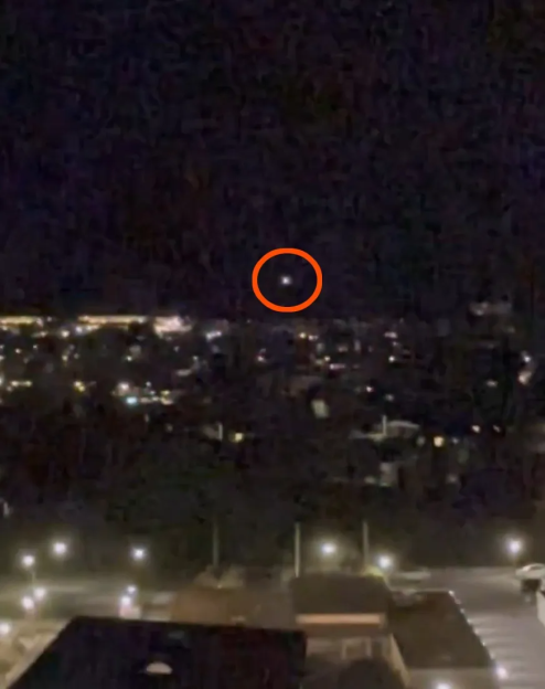 A bright light, possibly a UFO, circled in red, hovering over a city at night.