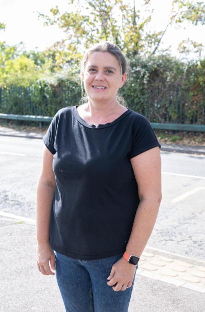 Zoe Cairns, a woman with dark hair pulled back, wearing a black t-shirt, blue jeans, and an orange smartwatch, standing outdoors and smiling at the camera.