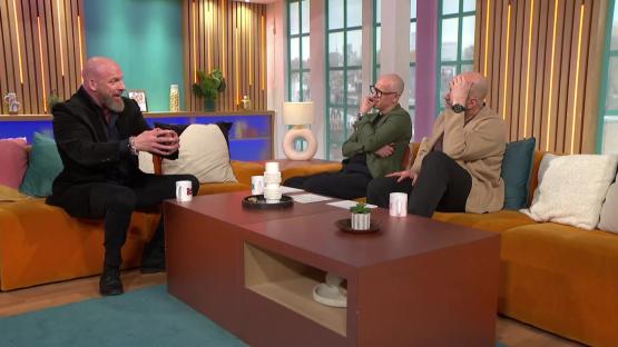 Three men sitting on an orange couch in a television studio, talking.