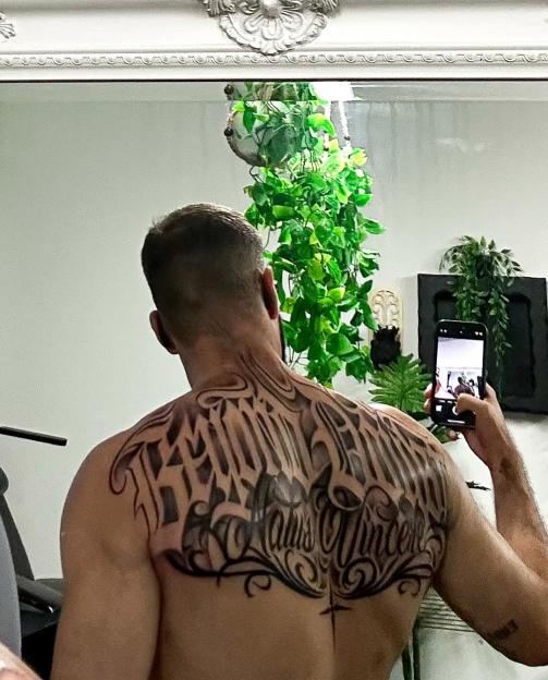 The back of a man with a large tattoo across his shoulders and upper back, taking a picture of himself in a mirror with a phone.