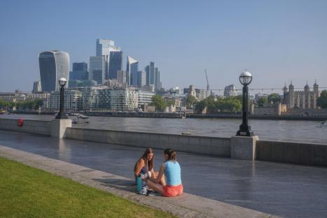 Two women relaxing by the London River, with the city skyline in the background.