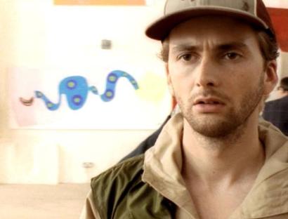 David Tennant in a beige jacket, looking pensive, in front of an abstract painting.