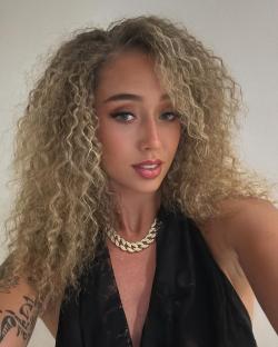 Close-up of a woman with curly blonde hair wearing a black top and a gold necklace.