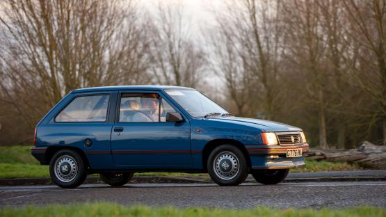 A blue 1989 Vauxhall Nova driving down a road.