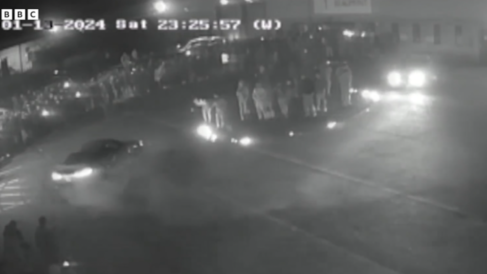 Surveillance footage shows a car doing donuts in the middle of a street at night, with a crowd of people watching on both sides of the street.