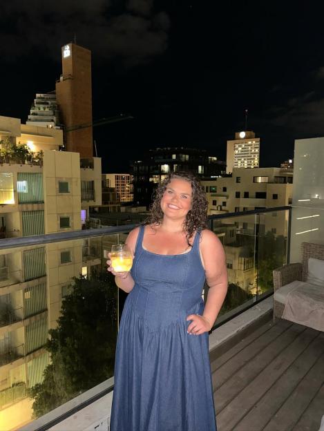 Amy Tapper in a denim dress holding a glass, standing on a balcony at night with city buildings in the background.