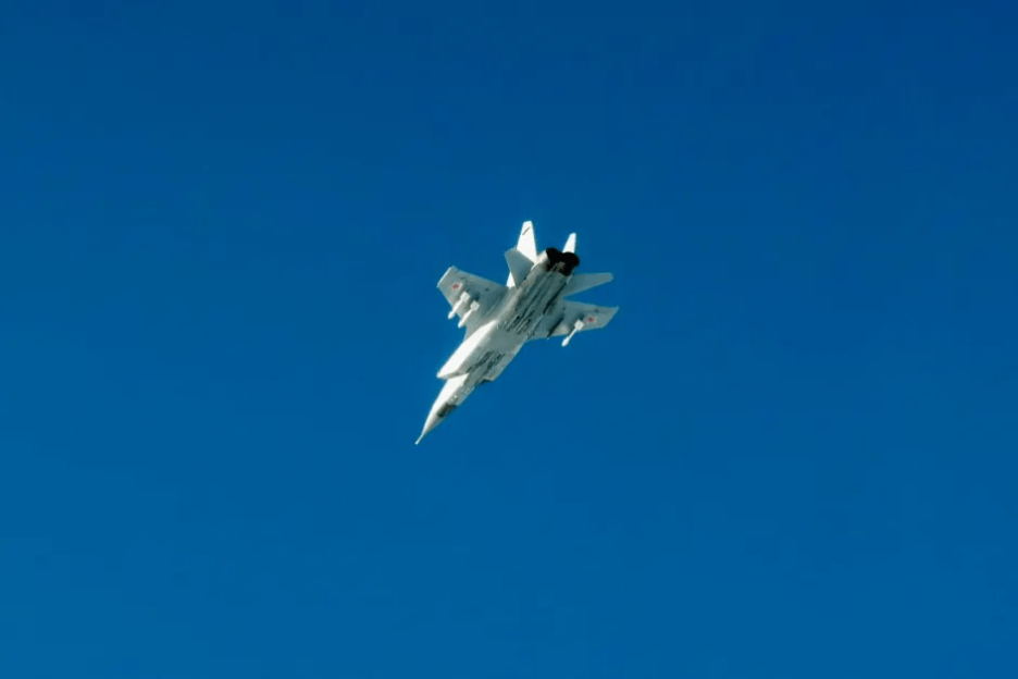 A white MiG-31 fighter jet flying against a clear blue sky.