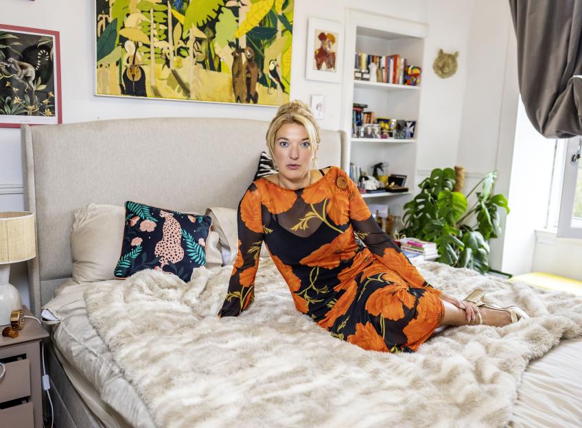 Journalist Lucy Holden in a black and orange floral dress, sitting on a bed.