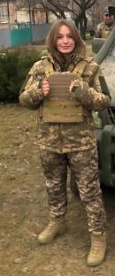 A female Ukrainian soldier with a prosthetic leg stands in front of a military vehicle.