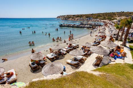 Beach scene in Cesme, Izmir, Turkey, with many people swimming and relaxing under umbrellas.