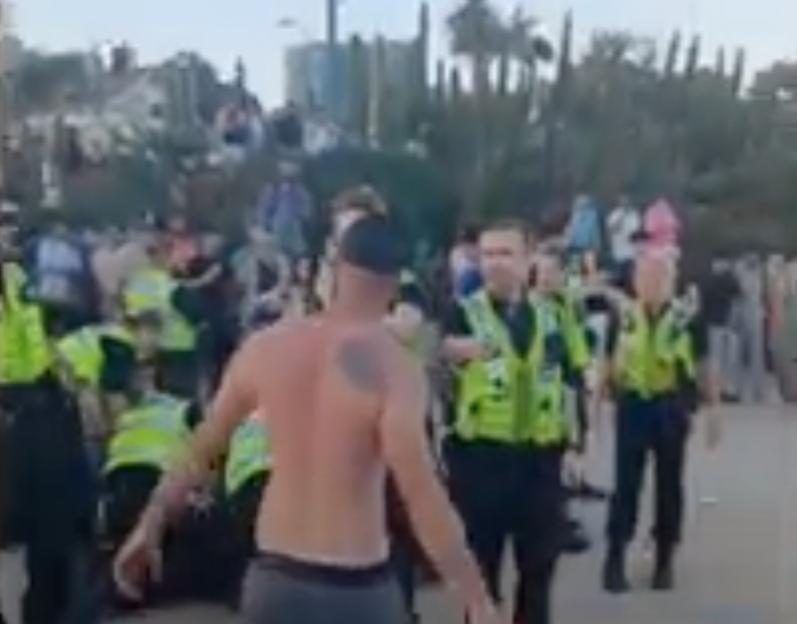 A shirtless man confronting police officers during a mass brawl on a beach.