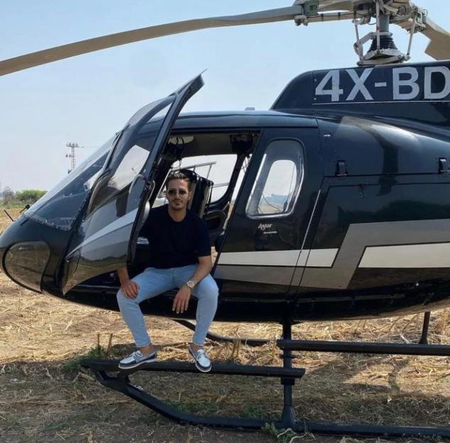 Simon Leviev sitting on the landing skid of a black helicopter with the door open.