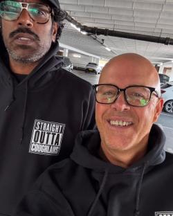 Romesh Ranganathan and another man wearing black hoodies in a parking garage.