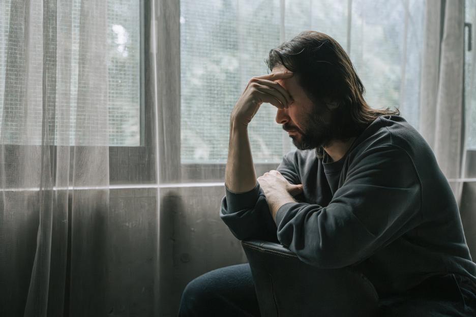 A contemplative adult man sitting alone indoors with head resting on his hand, looking distressed or emotionally overwhelmed. Captured in natural window light, the scene conveys feelings of melancholy, anxiety, solitude, and introspection. Mental health, depression, burnout, emotional struggle, or personal reflection concept.