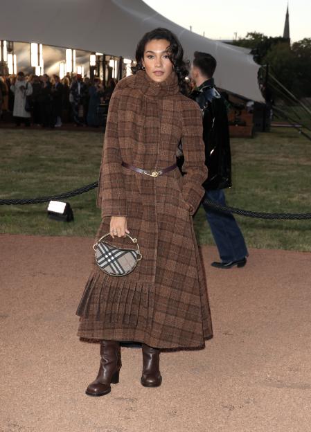 Olivia Dean wearing a brown plaid coat, brown boots, and carrying a Burberry crescent-shaped bag at the Burberry show.
