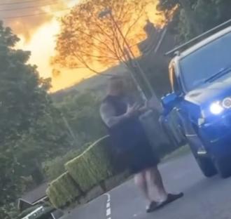 Screenshot of a man standing in the road next to a blue car at sunset.