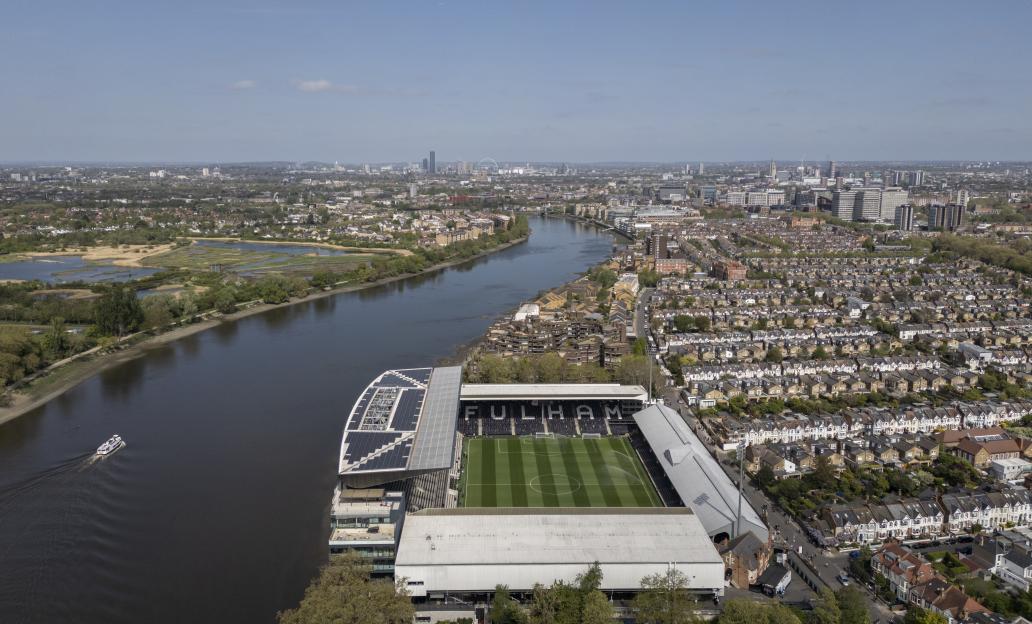 Aerial view of Craven Cottage stadium and River Thames, with a city skyline in the background.