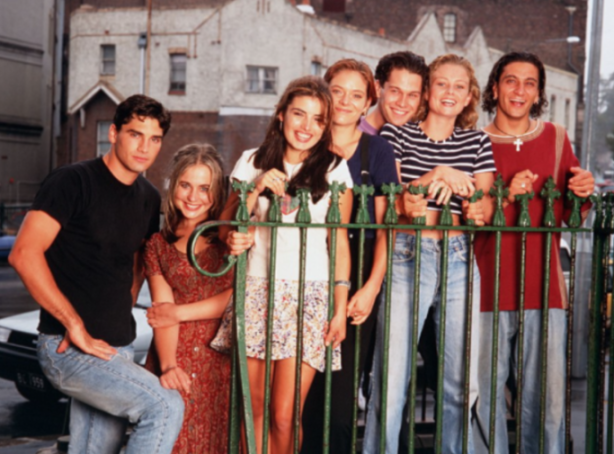 Six young adults posing behind a green fence in an urban setting.